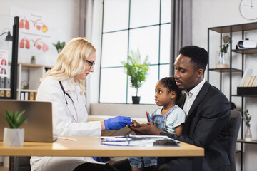 Obraz premium Caucasian doctor checking oxygen saturation of little african girl using modern pulse oximeter. Cute patient sitting on knees af her caring father. Family visiting doctor for controlling health.