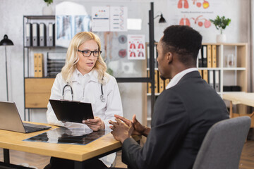 Caucasian female doctor in lab coat and eyeglasses talking with male patient at hospital office....