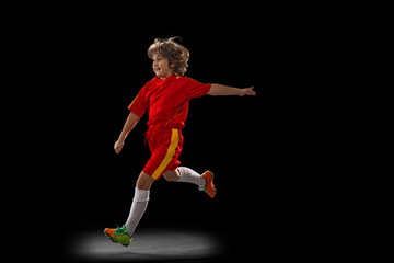 One little male football soccer player, boy training with football ball isolated on dark studio background. Concept of sport, game, hobby