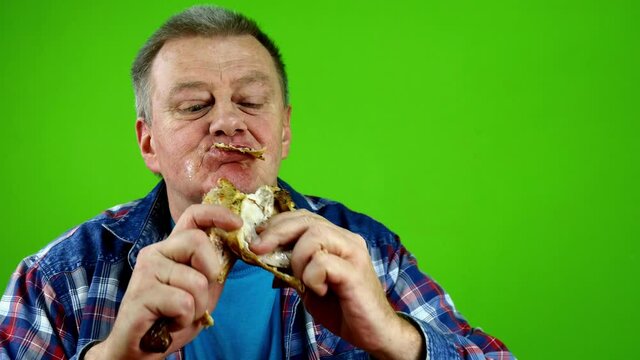 Hungry senior adult caucasian male with pleasure eating fried chicken leg, gnawing teeth into meat and wiping his mouth with his hand. Green screen. Chrome key. Close-up.