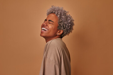 Fototapeta premium Overemotive African American woman laughs loudly hears funny joke or story, being overjoyed raises palms with satisfaction, dressed in beige t shirt, amused by friend. People and emotions concept.