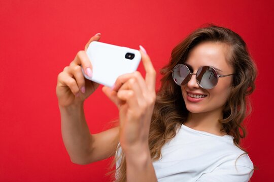 Closeup photo of amazing beautiful young blonde woman holding mobile phone taking selfie photo using smartphone camera wearing sunglasses isolated over colorful wall background looking at device