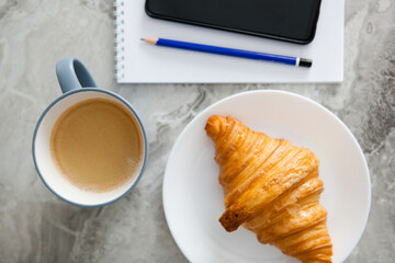 business breakfast in a light key, coffee and a croissant, a white notebook, a black phone and a blue pencil