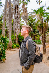 A young man with a backpack walks through a rainforest with trees with huge roots in Balboa Park, San Diego