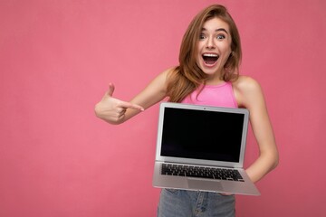 Naklejka premium Beautiful amazed shocked young woman holding computer laptop with empty monitor display wearing pink crop top looking at camera showing at netbook isolated on pink background