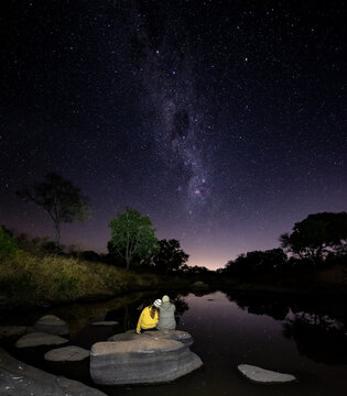 Mand and woman sit on a boulder together next to a river watching the milkway