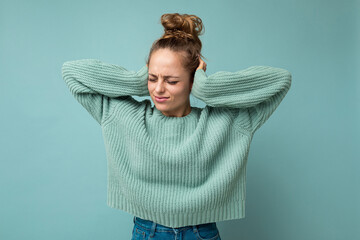 Portrait of young emotional sad dissatisfied attractive blonde woman with sincere emotions wearing casual blue jersey isolated over blue background with empty space and covering ears