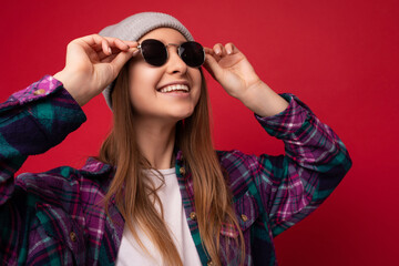 Closeup photo of attractive smiling positive young dark blonde woman isolated over red background wearing colourful stylish shirt grey hat and sunglasses looking up