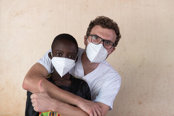 White doctor with thumb up hugging a litle black boy after medical examination, both wearing face...