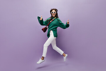Fototapeta na wymiar Attractive woman in stylish outfit moves and shows thumbs up. Pretty brunette girl on white pants and green sweater walks in good mood on isolated purple background.