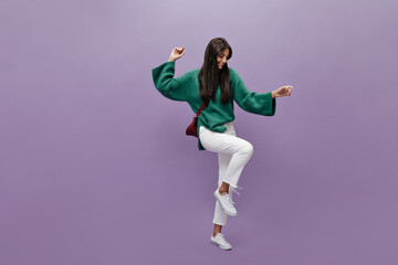 Fototapeta na wymiar Long-haired brunette woman in green knitted sweater dances on purple background. Happy girl in white pants moves on isolated.