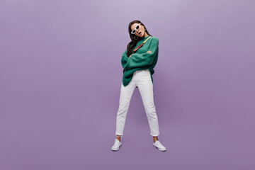 Fototapeta na wymiar Serious brunette woman in white pants and oversized green sweater poses on purple background. Full-length portrait of charming girl in sunglasses on isolated.