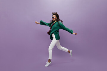 Fototapeta na wymiar Pretty tanned woman in green sweater and white pants moves on isolated. Charming young girl in sunglasses walks and jumps on purple background.
