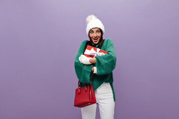 Surprised tanned woman in green sweater and white pants holds red gift box with white ribbon. Pretty brunette girl in knitted hat and gloves smiles on purple background.