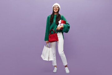 Happy young woman in woolen oversized green sweater, knitted hat, white pants and eyeglasses holds red gift box and shopping bags on purple background.
