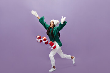 Young woman drops red gift boxes on purple background. Tanned charming girl in green sweater and white pants moves on isolated.