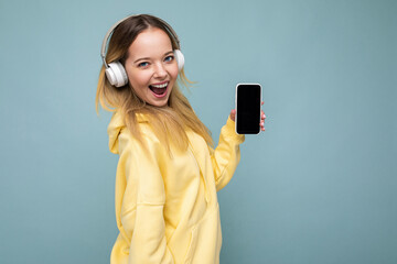 Side-profile beautiful positive young blonde woman wearing yellow stylish hoodie isolated on blue...