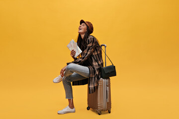 Cool girl holds tickets and sits on suit bag. Portrait of brunette long-haired woman in beret, jeans and coat smiling and posing on yellow background.