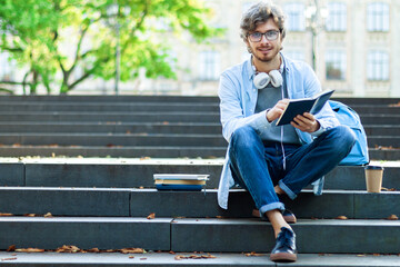 Obraz premium Happy university student is reading book while sitting on steps in campus. Caucasian young man is preparing for lessons at college, copy space area for your text message, content. Back to high school