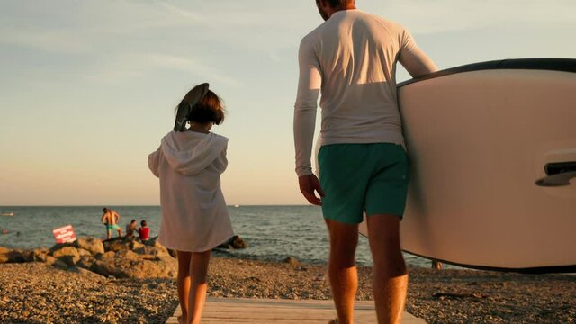 Surfing. Father with sup board and daughter with paddle are walking along the beach. Pan shot, Slow motion.
