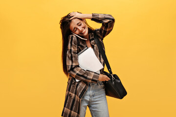 Tanned happy woman touches her dark hair and laughs. Charming girl in oversized jacket holds notebooks and talks on beige phone on isolated.
