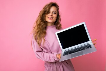 Fototapeta premium Photo of beautiful smiling happy blond curly young woman lookind at camera holding computer laptop with empty monitor screen with mock up and copy space wearing pink hoodie isolated over pink wall