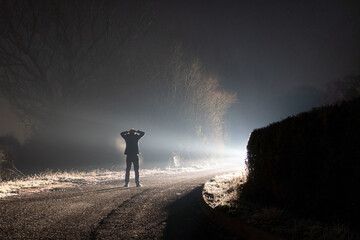 A figure standing on a road surrendering with his hands on his head. On a foggy winters night