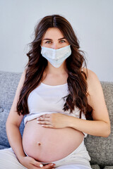 Young beautiful pregnant woman in medical surgical mask and white t-shirt quarantined at home by the window. Coronavirus, virus.