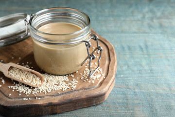 Jar of tasty sesame paste, seeds and scoop on light blue wooden table, space for text