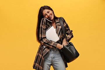 Cheerful woman in jeans and oversized jacket laughing on isolated. Pretty girl talks on phone, holds notebooks and black handbag on isolated yellow background.