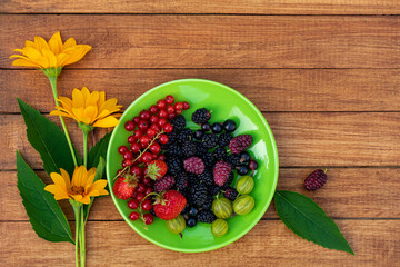 Berry mix - currants, gooseberries, mulberries, strawberries on a green saucer with yellow flowers on a wooden background with space for design. Summer concept.