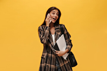 Happy woman in oversized stylish jacket talks on phone on isolated. Brunette long-haired girl holds notebooks on yellow isolated background.