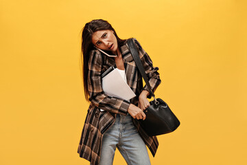 Obraz na płótnie Canvas Dissatisfied sad brunette woman talks on phone. Long-haired girl in oversized jacket holds handbag and notebooks on isolated yellow background.