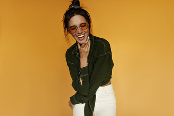 Charming woman in white pants and dark green shirt laughs on isolated. Brunette girl in orange sunglasses smiles and poses on yellow background.