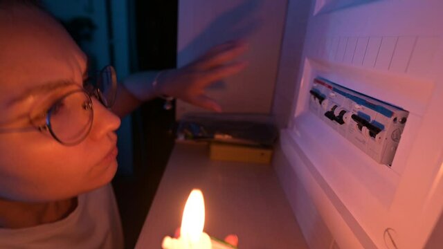 Young woman with a candle checks a row of circuit breakers in fuse box during a power outage in an area