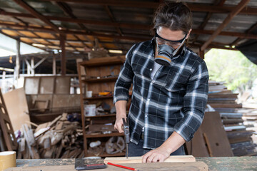 Male carpenter using hammering a nail into wooden board and wearing safety goggles and dust filter mask at the carpentry workshop. Joiner using hammer and nails fixes a wooden plank