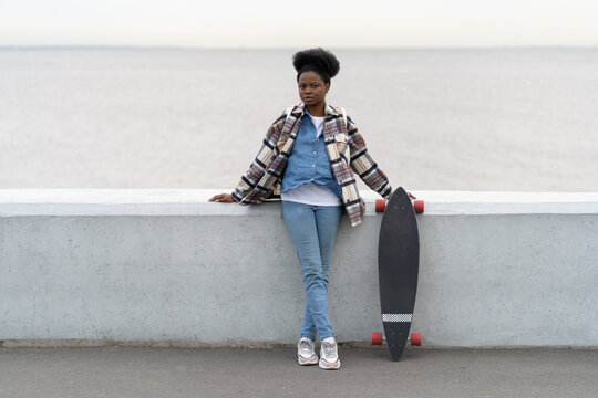 Young afro american woman with longboard over river stand meditating and recreating after skateboarding on promenade. Trendy multiracial african girl student alone outdoors. Urban lifestyle concept - Powered by Adobe