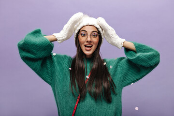 Tanned brunette woman in white knitted hat and gloves looks into camera on isolated. Pretty girl in eyeglasses and green sweater poses on purple background with snow flakes.