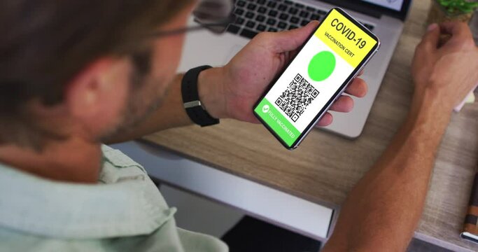 Man at desk holding smartphone with covid vaccination certificate and qr code on screen