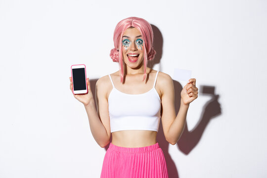 Image of excited party girl in pink wig and halloween outfit, showing credit card and smartphone screen, smiling happy over white background