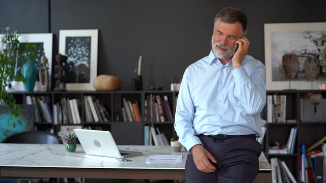 Smiling mature businessman holding smartphone standing in office. 60s aged manager ceo using cell phone mobile apps. Digital technology applications and solutions for business corporate development