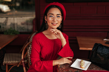 Tanned Asian woman in red beret, bright dress and eyeglasses smiles, sits in gorgeous cafe and looks into camera.