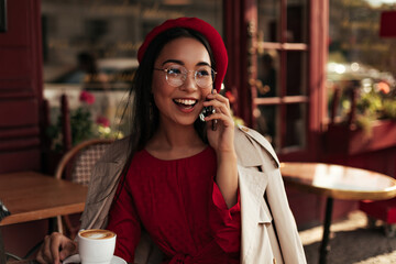 Cheerful tanned brunette woman in red beret, dress and beige trench coat smiles widely and talks on phone in street cafe.