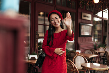 Cheerful lady in red beret and bright dress waves hand in greeting. Pretty woman in eyeglasses and stylish outfit smiles and poses in street cafe.