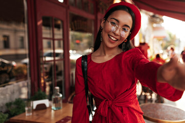 Pretty brunette tanned woman in stylish beret, red dress and eyeglasses smiles and takes selfie in cozy wooden street cafe.