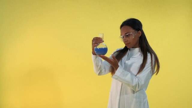 Black female scientist in lab coat looking on flask with experimental liquid on yellow background. She also has safety glasses on eyes.