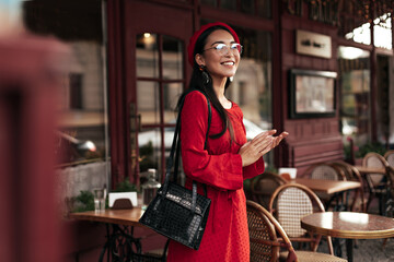 Brunette attractive woman in stylish beret, red dress and eyeglasses smiles widely and poses in cozy street cafe.
