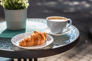 Breakfast with croissant and a cup of cappuccino on the table early sunshine morning in a street cafe.