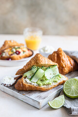 Croissant sandwich with ricotta or cream cheese, mashed avocado and cucumber on wooden cutting board. Delicious brunch or breakfast.