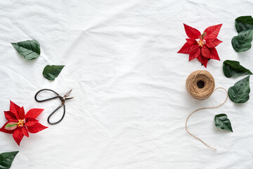Christmas flat lay with copy-space. Red poinsettia, Xmas top view on off white, ivory textile background, text space. Hemp cord, scissors, red and dark green poinsettia leaves .